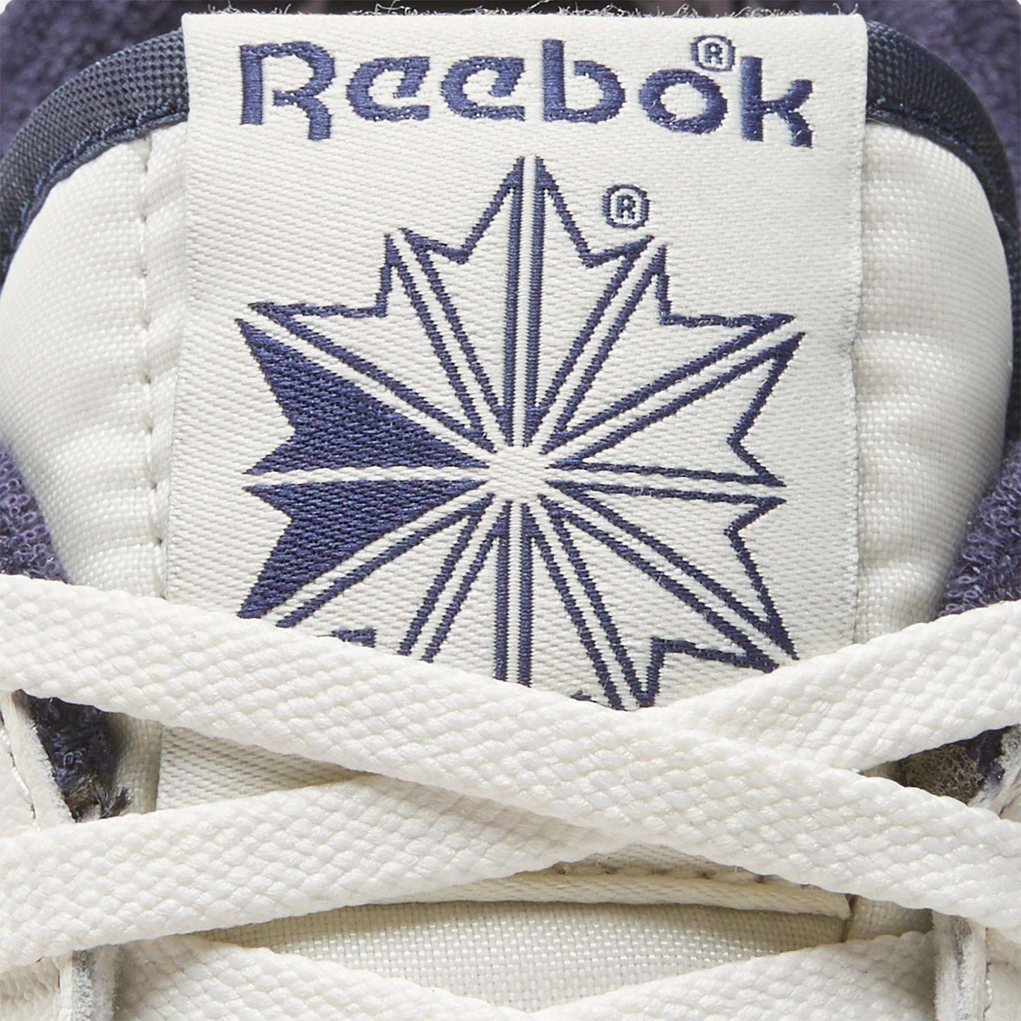 Close-up of the Men's Club C Golf Shoes by Reebok: a white tongue with blue Reebok logo and starburst, partially covered by laces. Crafted from water-resistant leather for lasting comfort on the course.
