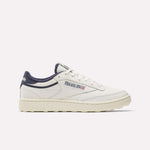 The Reebok Men's Club C Golf Shoes are white with navy blue accents, a beige spikeless outsole, and feature the Reebok logo with a small Union Jack on the side, displayed in a side profile on a plain white background.