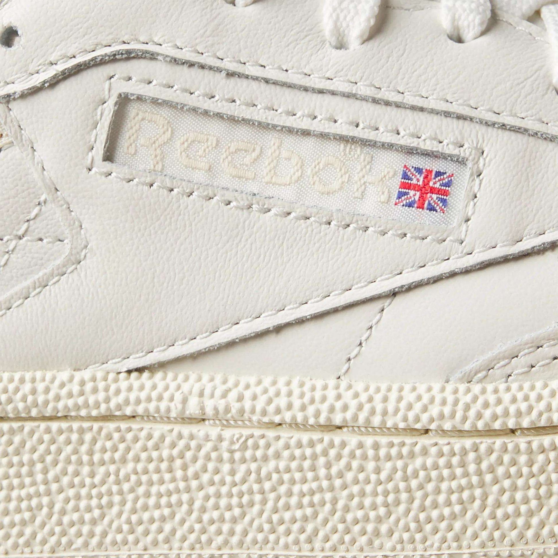 Close-up of Women's Club C Golf Shoes by Reebok in white water-resistant leather, featuring the Reebok logo, a small Union Jack flag on the side, fine stitching, and a spikeless, textured golf shoe-inspired sole.