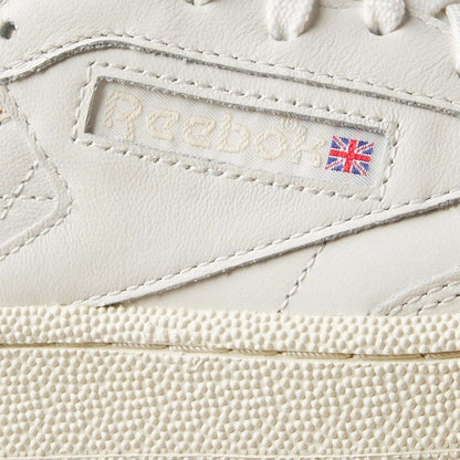 Close-up of Women's Club C Golf Shoes by Reebok in white water-resistant leather, featuring the Reebok logo, a small Union Jack flag on the side, fine stitching, and a spikeless, textured golf shoe-inspired sole.