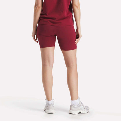 A person stands with their back to the camera, wearing a matching dark red t-shirt and Reebok Women's Small Logo Cycle Shorts, paired with white socks and sneakers, against a plain white background.
