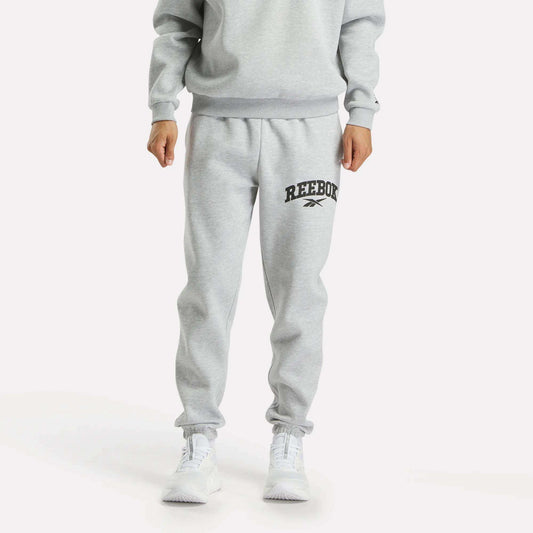A person models Reebok Men's Joggers in light gray with a black logo on the left thigh, styled with a matching sweatshirt and white sneakers against a plain white background.