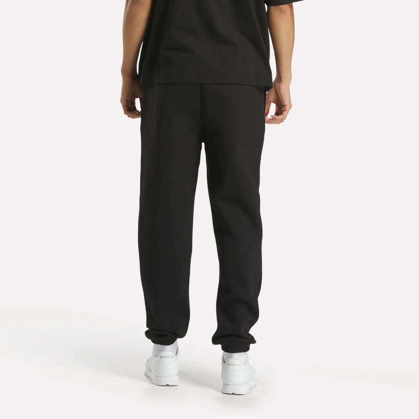A person stands facing away from the camera against a plain white background, wearing a loose black T-shirt, Reebok Men's Joggers, and white sneakers.