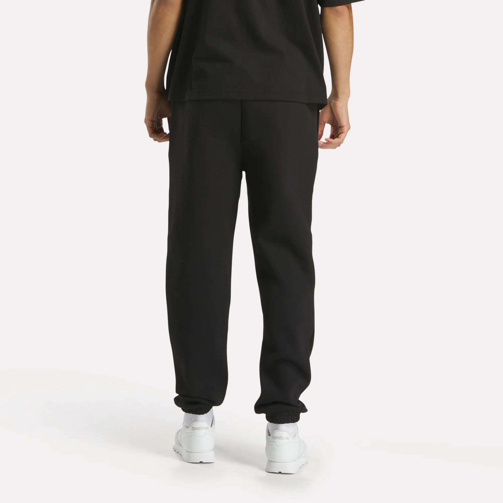 A person stands facing away from the camera against a plain white background, wearing a loose black T-shirt, Reebok Men's Joggers, and white sneakers.