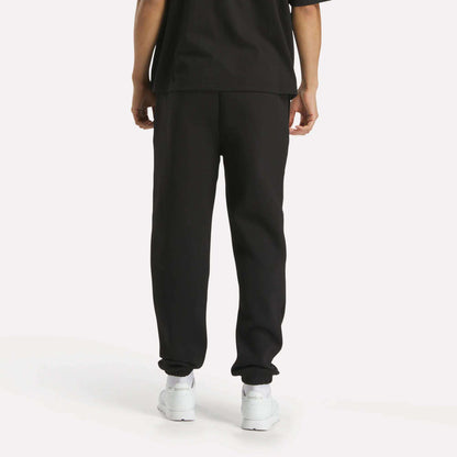 A person stands facing away from the camera against a plain white background, wearing a loose black T-shirt, Reebok Men's Joggers, and white sneakers.