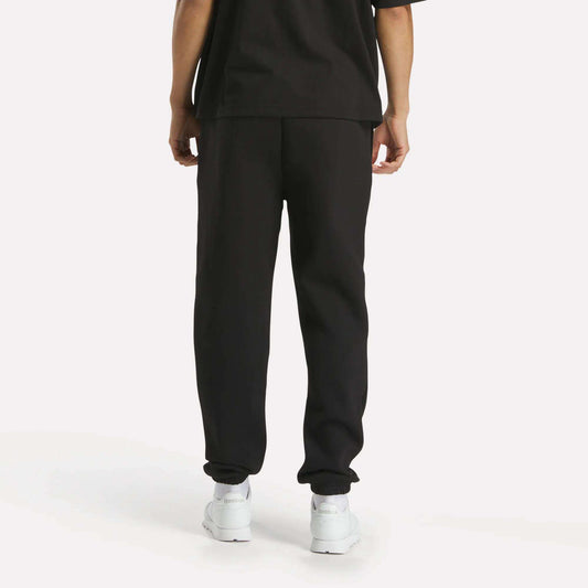 A person stands facing away from the camera against a plain white background, wearing a loose black T-shirt, Reebok Men's Joggers, and white sneakers.