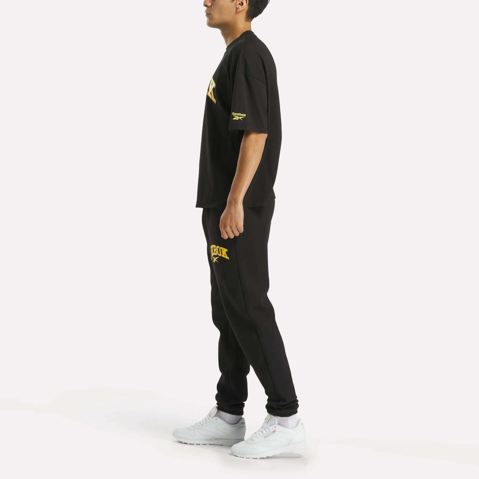 A person in profile wears a black T-shirt and Reebok Men's Joggers, black with yellow text, plus white sneakers, shown against a plain white background.