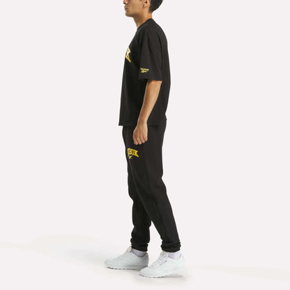 A person in profile wears a black T-shirt and Reebok Men's Joggers, black with yellow text, plus white sneakers, shown against a plain white background.