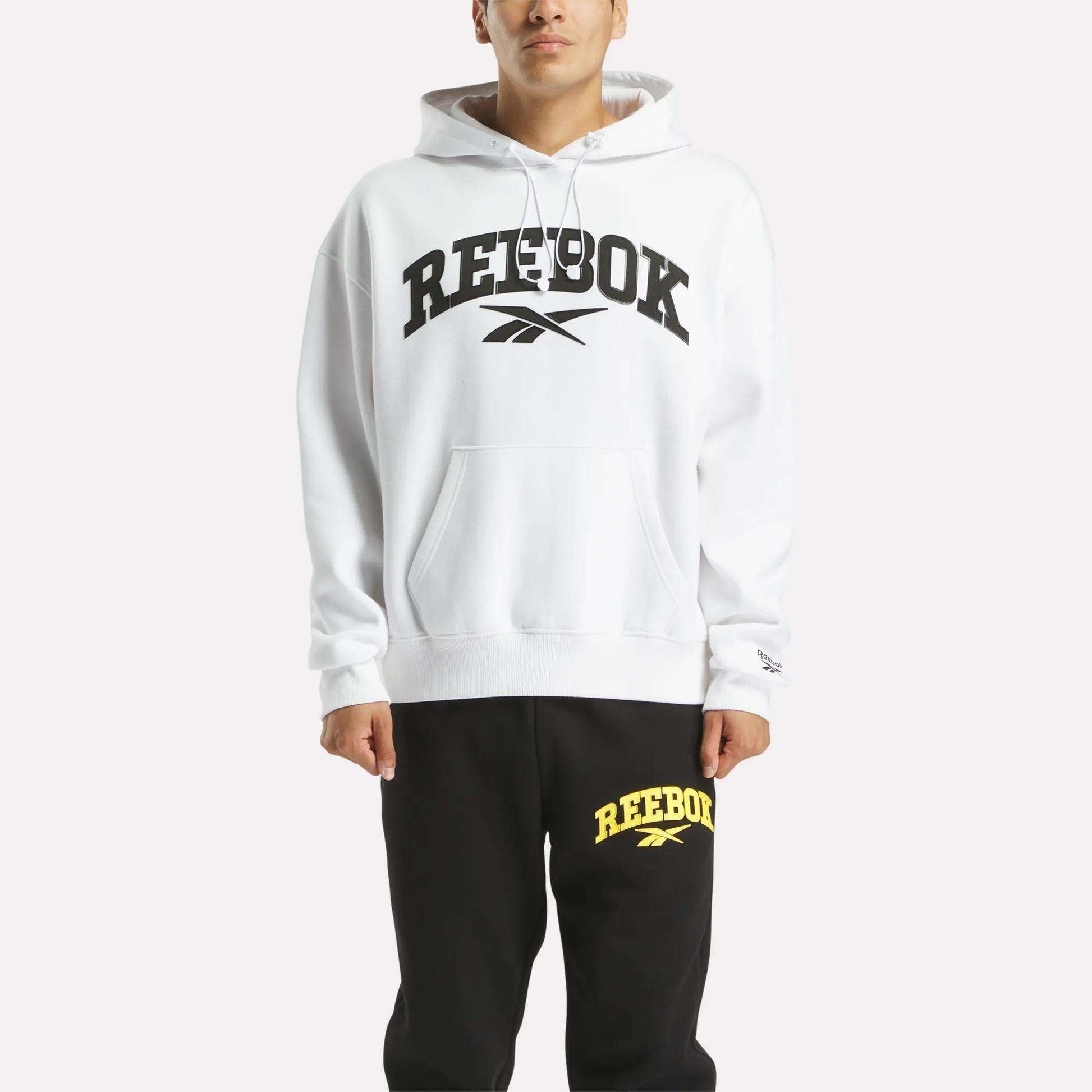 A person wears a soft white Reebok Men's Hoodie with a bold black logo, paired with black sweatpants featuring a yellow Reebok logo, standing against a plain light background.