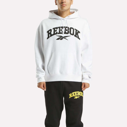 A person wears a soft white Reebok Men's Hoodie with a bold black logo, paired with black sweatpants featuring a yellow Reebok logo, standing against a plain light background.