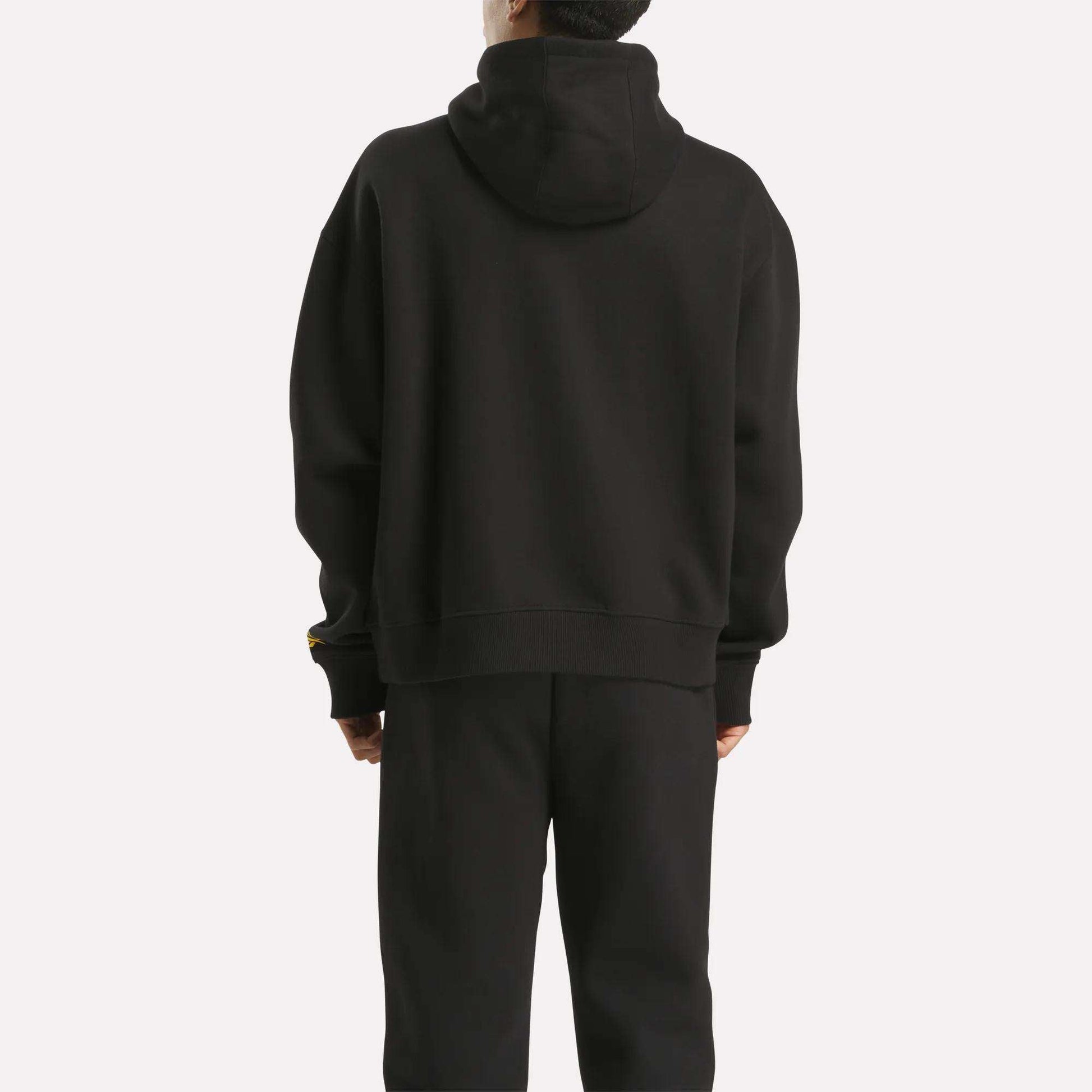 A person stands with their back to the camera, wearing a Men's Hoodie by Reebok in black, paired with matching black pants against a plain white background.