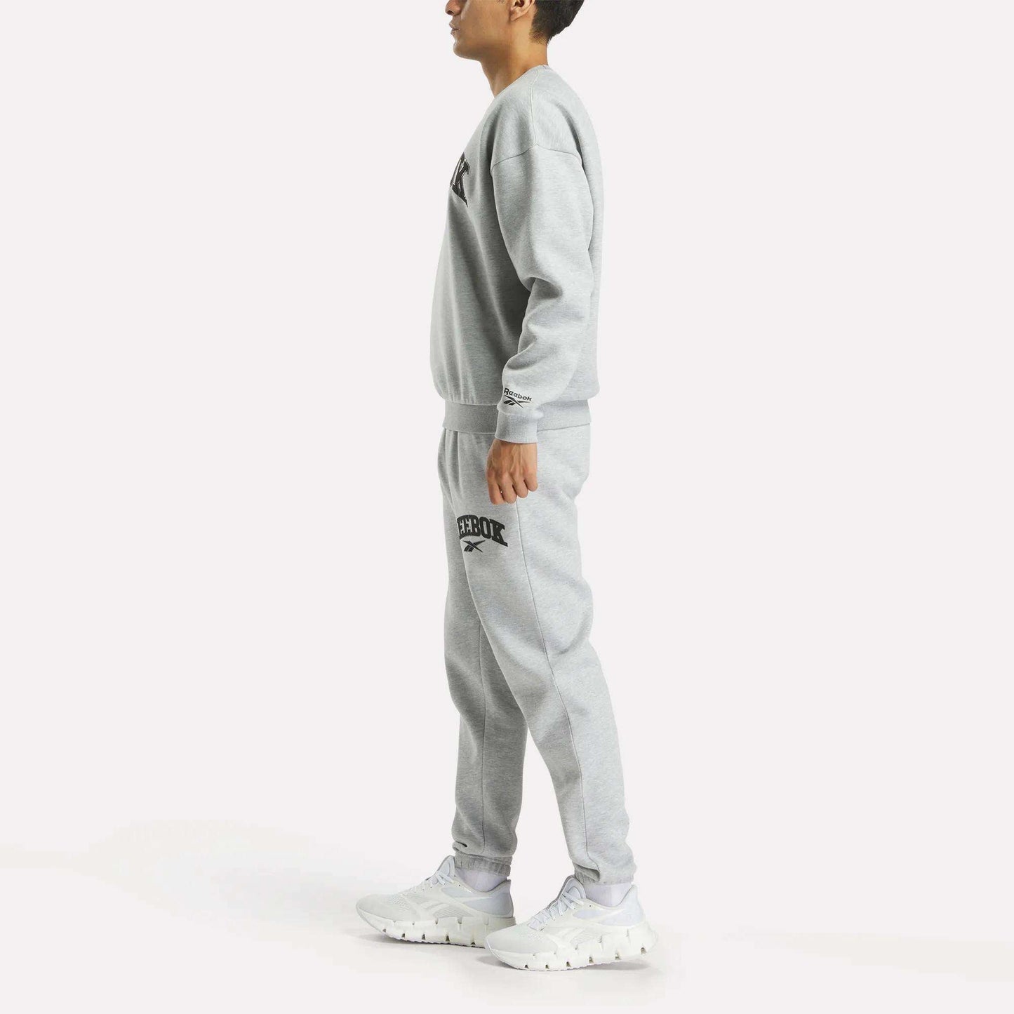 A person stands sideways in a matching grey Reebok Men's Cottle Sweatshirt and joggers with black logos, paired with white sneakers, against a plain white background.