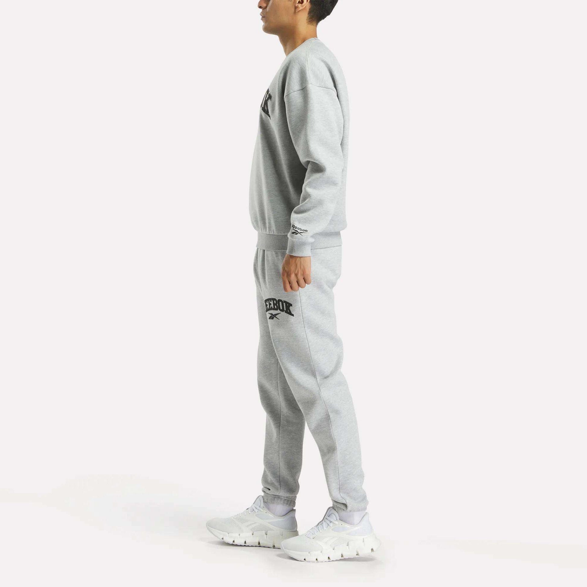 A person stands sideways in a matching grey Reebok Men's Cottle Sweatshirt and joggers with black logos, paired with white sneakers, against a plain white background.