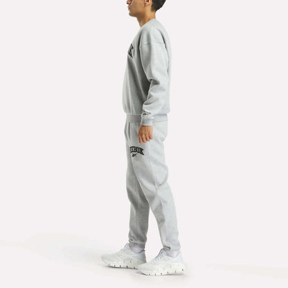 A person stands sideways in a matching grey Reebok Men's Cottle Sweatshirt and joggers with black logos, paired with white sneakers, against a plain white background.