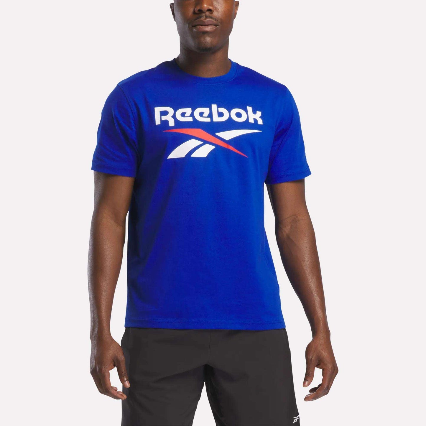 A person wearing the Reebok Men's Logo Crewneck T-Shirt in bright blue, featuring a large white and red vector logo across the chest, paired with black shorts, stands against a plain white background.