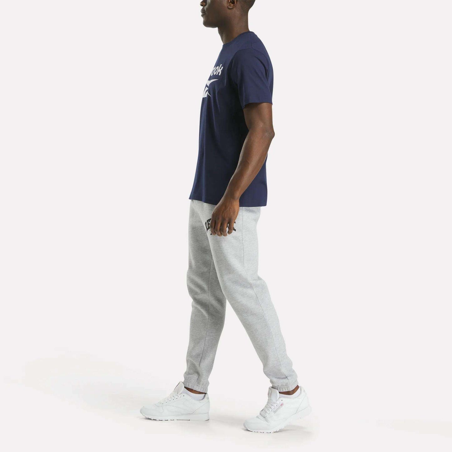 A man stands sideways against a plain white background wearing a Reebok Men's Big Logo Crewneck Short Sleeve Tee in navy blue, light gray jogger pants, and white sneakers.