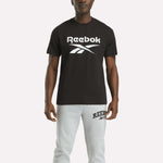 A person faces forward against a plain white background, wearing the Reebok Men's Big Logo Crewneck Short Sleeve Tee in black with a large white logo and light gray Reebok sweatpants.