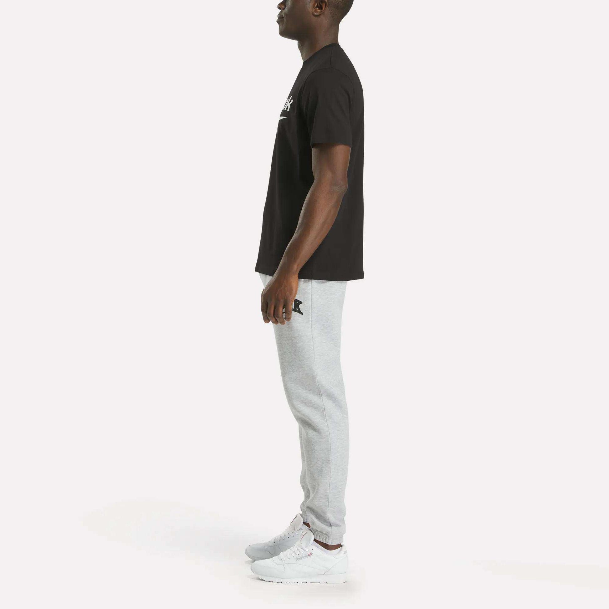 A man stands in profile against a plain white background, wearing Reebok's Men's Big Logo Crewneck Short Sleeve Tee, paired with light gray jogger pants and white sneakers. The tee features a bold Reebok logo on the chest.