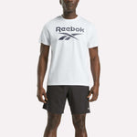 A person stands against a plain light background wearing a Reebok Men's Big Logo Crewneck Short Sleeve Tee in white and black athletic shorts.