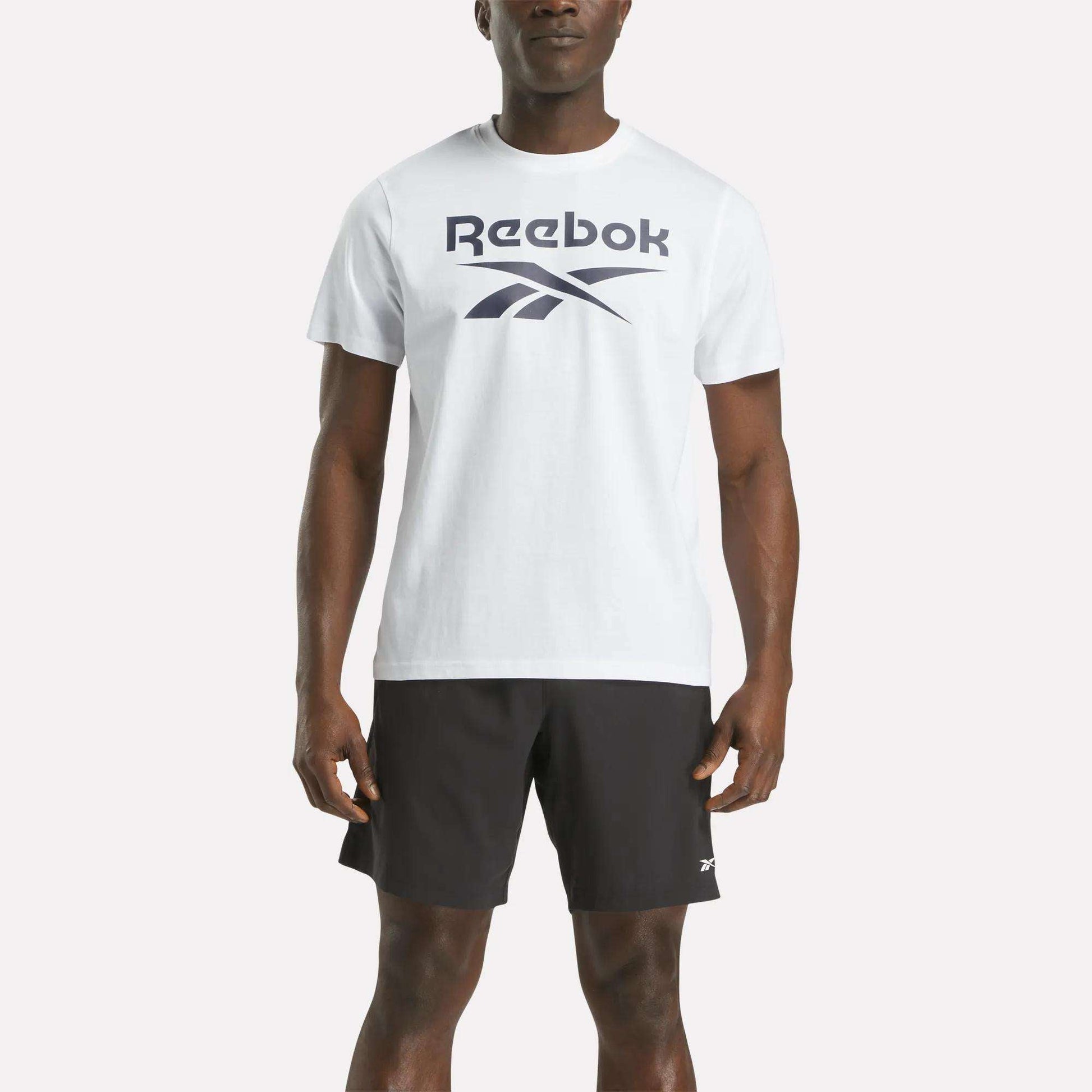 A person stands against a plain light background wearing a Reebok Men's Big Logo Crewneck Short Sleeve Tee in white and black athletic shorts.
