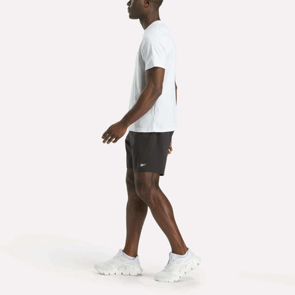 A man wears a Reebok Men's Big Logo Crewneck Short Sleeve Tee and black athletic shorts with white sneakers featuring a subtle Reebok logo, walking forward against a plain white background with his left arm slightly extended.