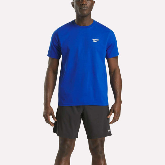 A man faces forward wearing a Reebok Men's Small Logo Crewneck Short Sleeve T-Shirt in blue and black Reebok shorts against a plain white background.