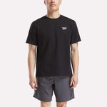 A man in a Reebok Men's Small Logo Crewneck Short Sleeve T-Shirt, black cotton jersey with a small white logo on the chest, paired with gray shorts, stands against a plain white background.