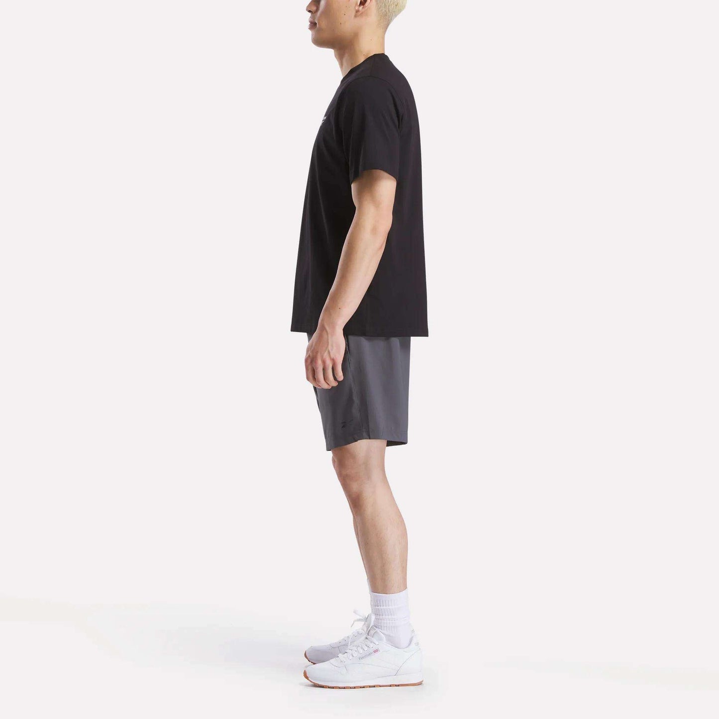 A man stands in profile against a white background, wearing a Reebok Men's Small Logo Crewneck Short Sleeve T-Shirt in black, gray shorts, white crew socks, and white athletic shoes.