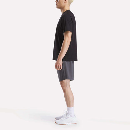 A man stands in profile against a white background, wearing a Reebok Men's Small Logo Crewneck Short Sleeve T-Shirt in black, gray shorts, white crew socks, and white athletic shoes.