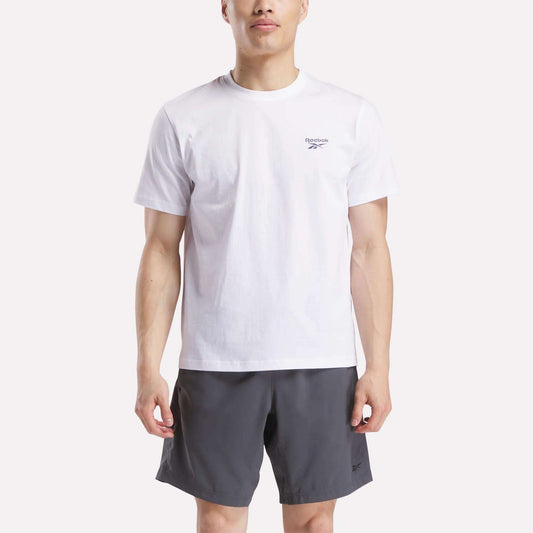 A man faces forward wearing a Reebok Men's Small Logo Crewneck Short Sleeve T-Shirt in white, made from soft cotton jersey, paired with dark gray shorts against a plain light background.