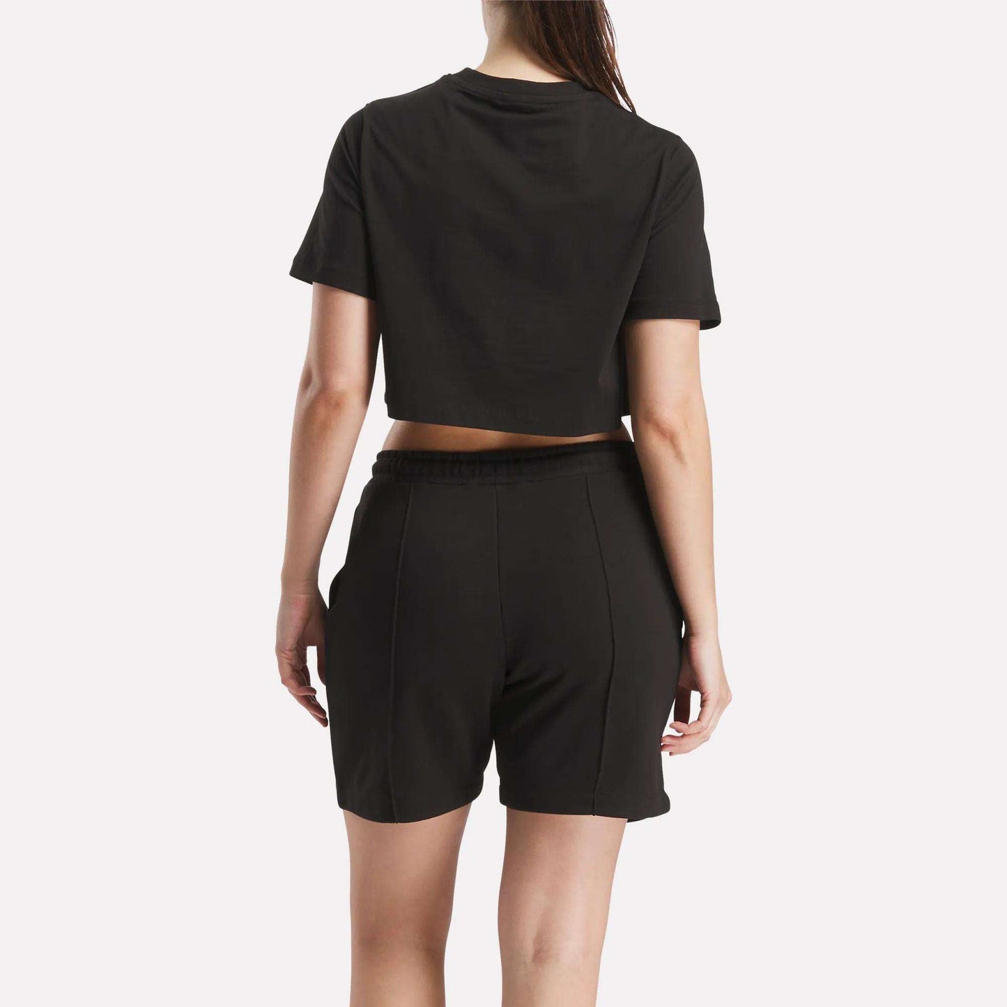 A woman faces away from the camera, wearing a Reebok Women's Two-Color Logo Crew Cropped Short Sleeve Tee in black with matching high-waisted shorts. Her right arm hangs by her side and her left arm is slightly bent.