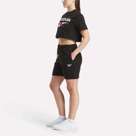 A woman stands in profile against a plain white background, wearing a Reebok Women's Two-Color Logo Crew Cropped Short Sleeve Tee in black, matching black shorts, white socks, and white sneakers.