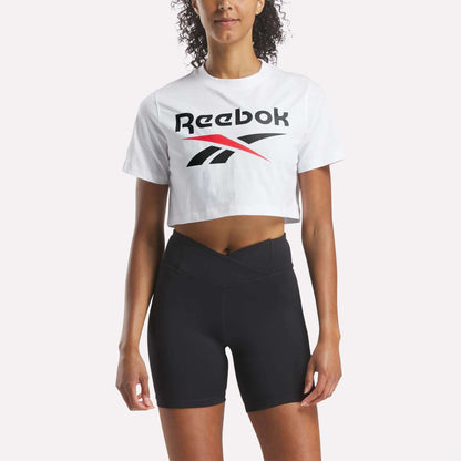 A woman faces forward against a plain light background, wearing a white Reebok Women's Two-Color Logo Crew Cropped Short Sleeve Tee and black high-waisted athletic shorts.