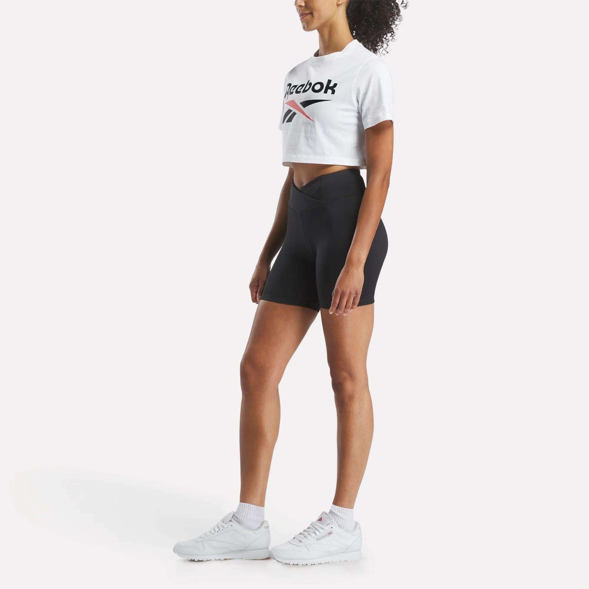 A woman in a Reebok Women's Two-Color Logo Crew Cropped Short Sleeve Tee, black high-waisted biker shorts, white socks, and sneakers stands in profile against a plain white background.