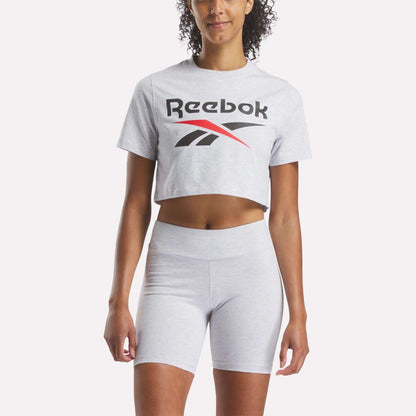 A woman wears a Reebok Women's Two-Color Logo Crew Cropped Short Sleeve Tee in light gray with matching high-waisted bike shorts, standing against a plain white background.