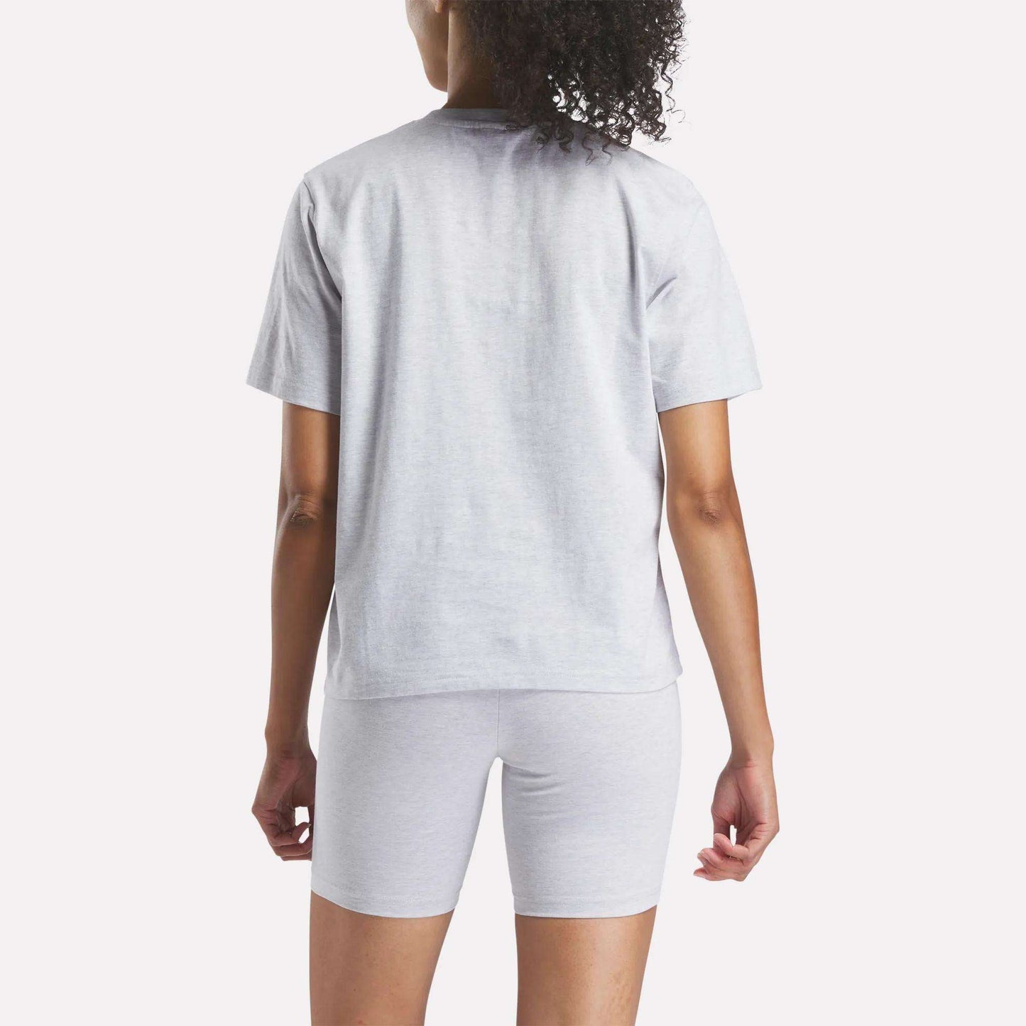 A person with curly hair is seen from behind wearing a light gray Reebok Women's Big Logo Oversized Crew Short Sleeve Tee against a plain white background.