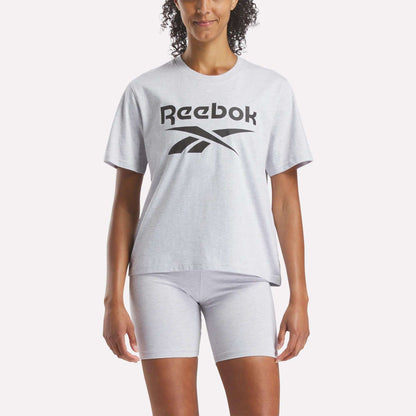 A person stands against a white background wearing a Reebok Women's Big Logo Oversized Crew Short Sleeve Tee in light gray with a black logo, paired with matching gray shorts.