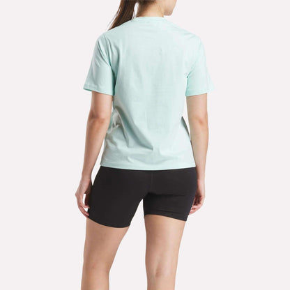 A person stands facing away, wearing a light blue Reebok Women's Two-Color Logo Crew Short Sleeve Tee and black shorts against a plain white background.
