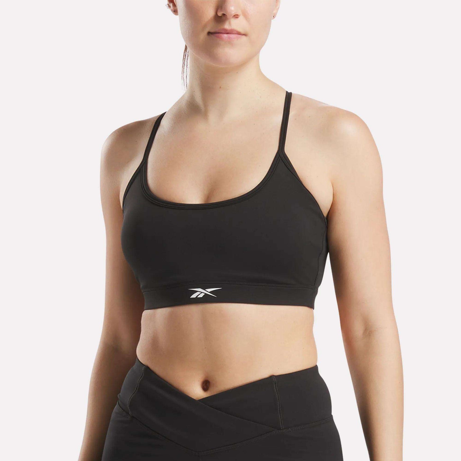 A woman wears the Reebok Women's Core Strappy Bra - Medium Support in black, paired with high-waisted athletic leggings, standing against a plain light background.