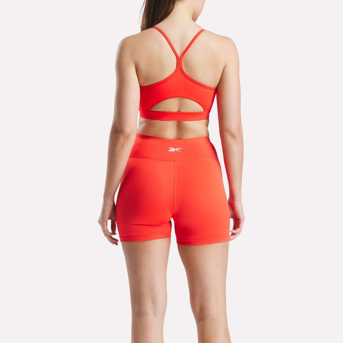 A woman, seen from behind, wears the Reebok Women's Core Strappy Bra - Medium Support and matching high-waisted shorts in bright red with logos, perfect for medium-impact workouts, set against a plain white background.