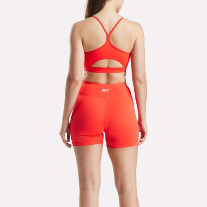 A woman, seen from behind, wears the Reebok Women's Core Strappy Bra - Medium Support and matching high-waisted shorts in bright red with logos, perfect for medium-impact workouts, set against a plain white background.