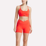 A woman wears the Reebok Women's Strappy Sports Bra and matching high-waisted shorts in bright orange against a white background. The quick-drying athletic set is perfect for medium-impact workouts and features a small Reebok logo on the bra.
