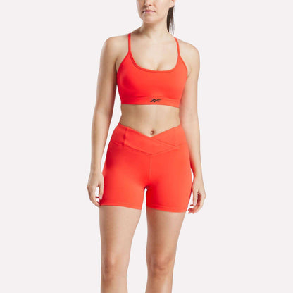 A woman wears the Reebok Women's Strappy Sports Bra and matching high-waisted shorts in bright orange against a white background. The quick-drying athletic set is perfect for medium-impact workouts and features a small Reebok logo on the bra.
