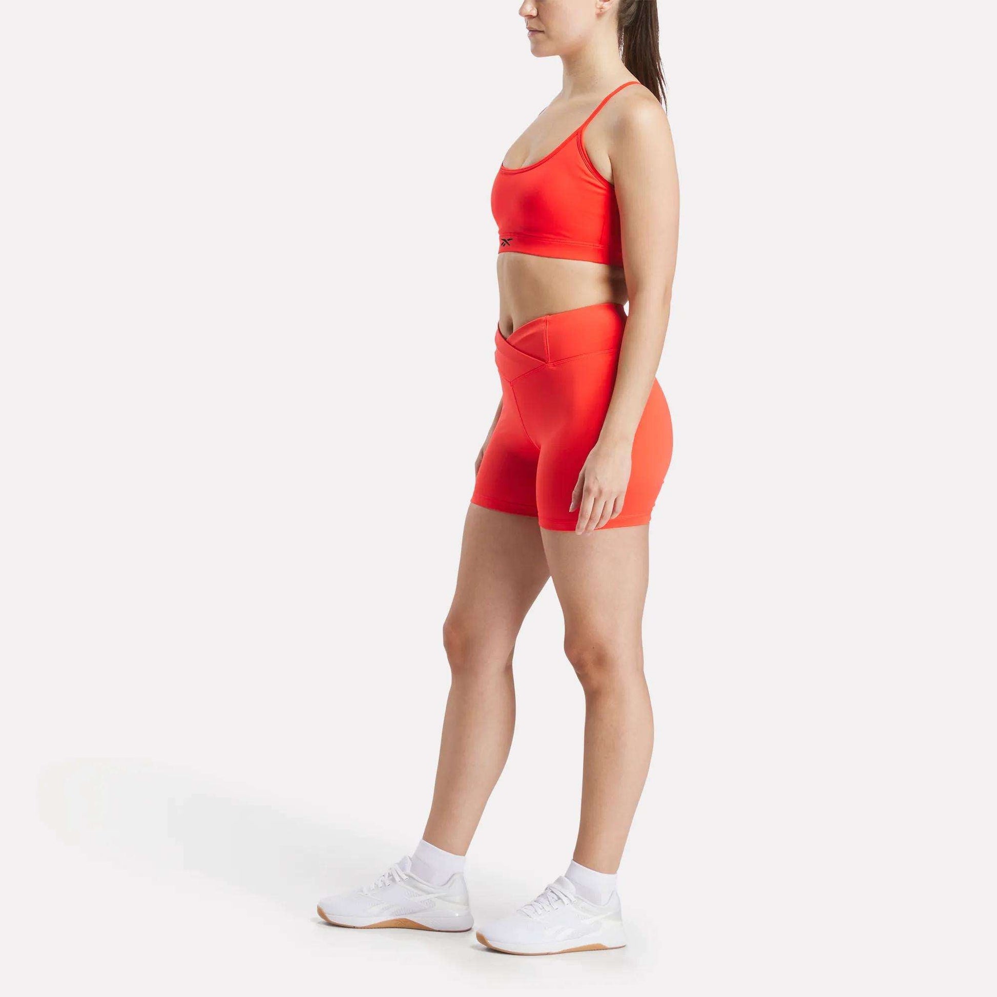 A woman stands in profile against a white background, wearing a bright orange Reebok Women's Strappy Sports Bra with matching high-waisted shorts, white socks, and sneakers—ideal for quick-drying comfort during medium-impact workouts.