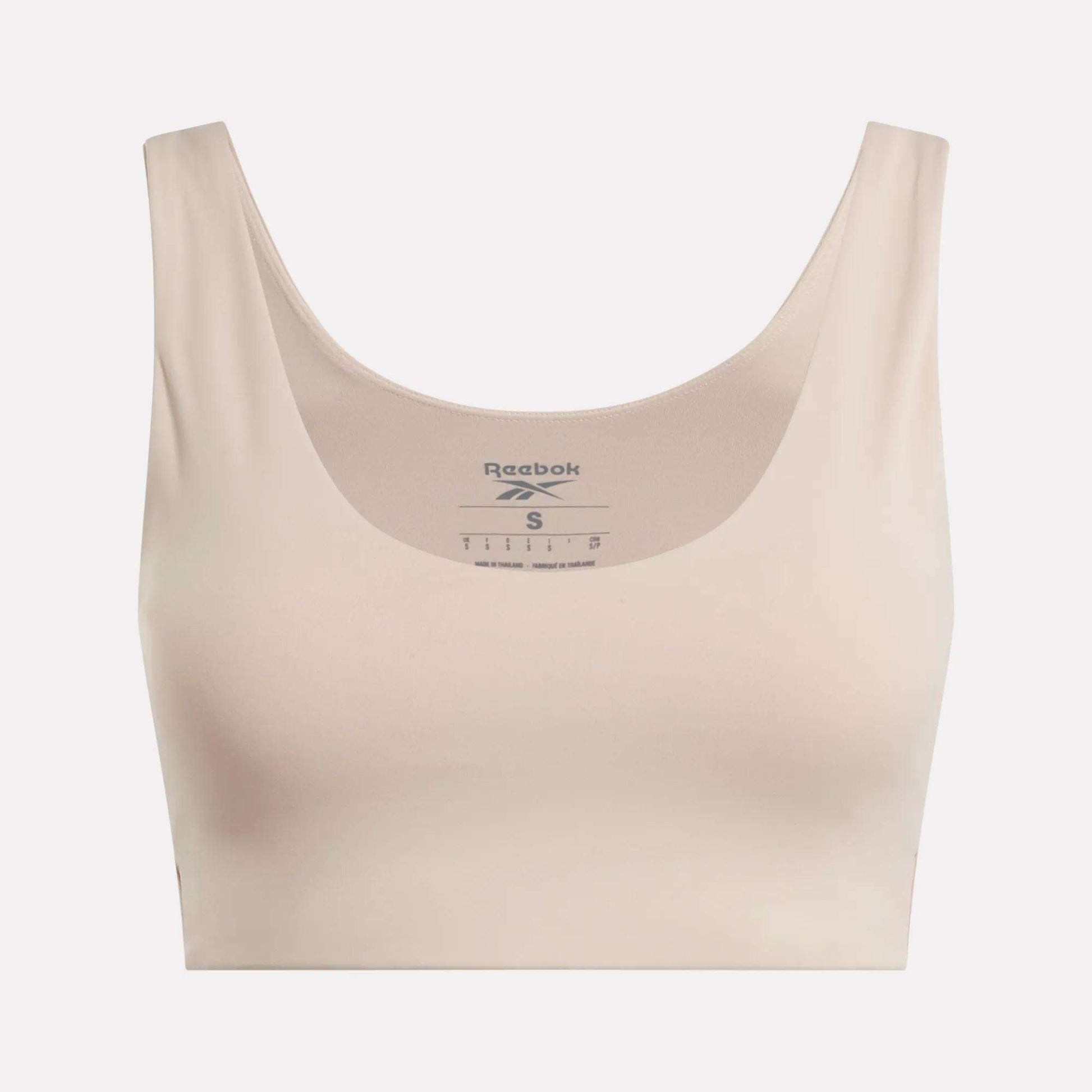 The Reebok Women's Yoga V-Neck Bra in beige features a scoop neckline and wide straps, offering medium-impact support for studio workouts. The Reebok logo and sizing appear inside the neckline on a plain light background.