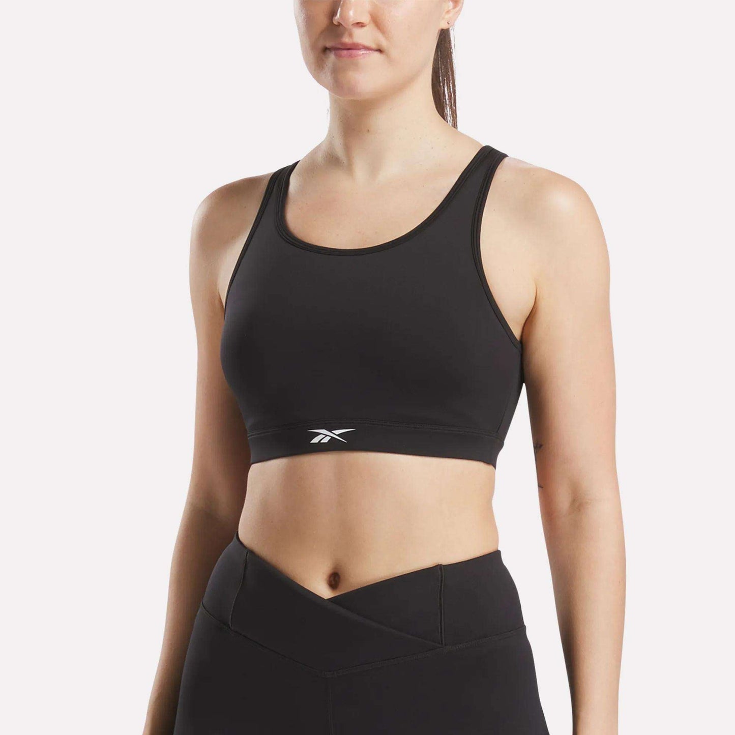 A woman in a black Reebok Women's Core High-Impact Bra with a small white logo and matching high-waisted leggings stands against a plain white background. The sweat-wicking bra offers support and style.