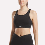 A woman in a black Reebok Women's Core High-Impact Bra with a small white logo and matching high-waisted leggings stands against a plain white background. The sweat-wicking bra offers support and style.