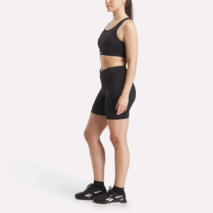 A woman stands sideways against a plain white background wearing the Reebok Women's Core High-Impact Bra, high-waisted black athletic shorts, and black sneakers with white accents.