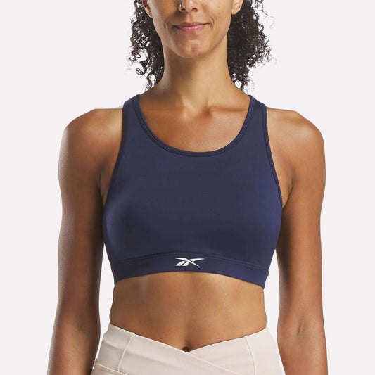 A woman wears the Reebok Women's Core High-Impact Bra in navy blue with light leggings, standing facing forward and smiling slightly against a plain white background.