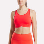 A woman wears the Reebok Women's Core High-Impact Bra, a bright orange racerback sports bra with a small white logo on the front, paired with matching high-waisted leggings against a plain, light background.